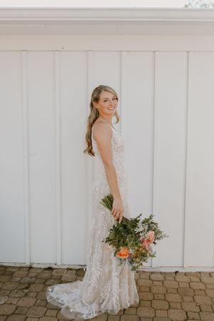 Bride in lace wedding dress poses with bouquet against white paneled wall.