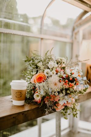 Beautiful wedding bouquet with coral roses and white flowers next to a coffee cup on a wooden ledge.