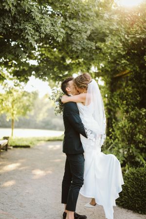 A romantic wedding portrait showing a couple embracing in golden sunlight under trees.