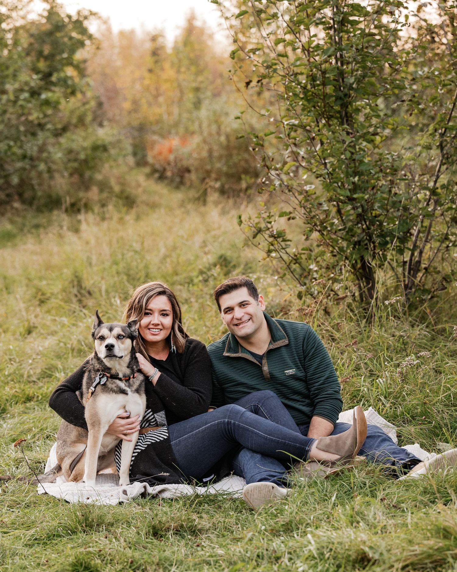 A husky dog and two people sitting together on a blanket in a grassy autumn field at sunset.