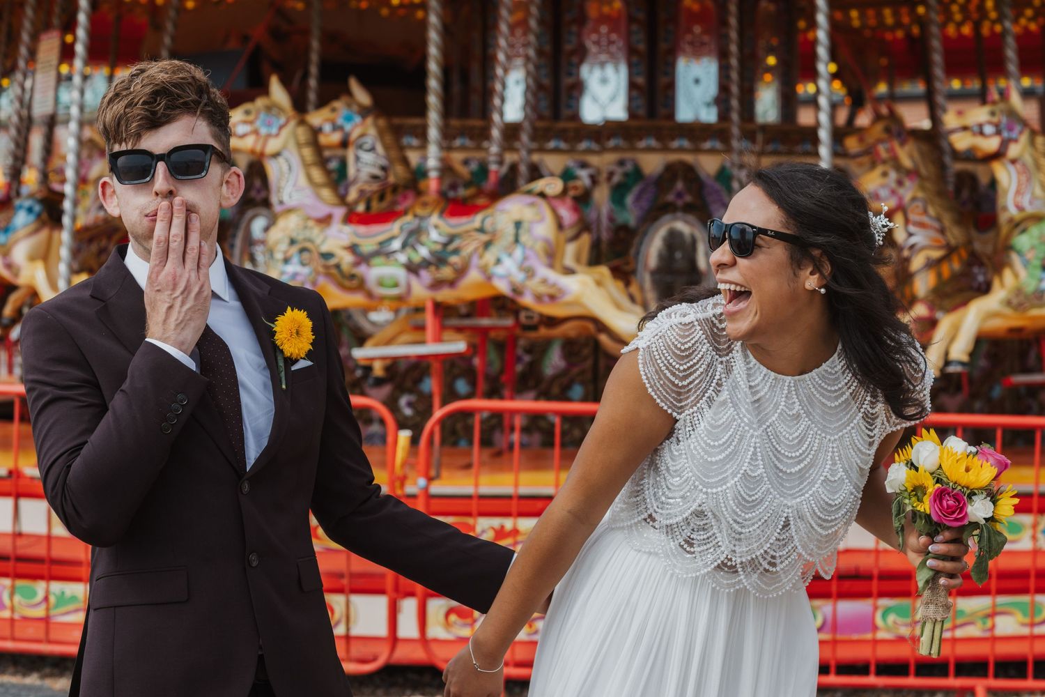 Simon and Natasha in their wedding attire laughing and having fun in front of a carousel on Brighton Seafront