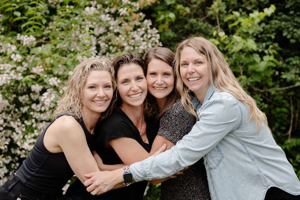 Four women posing for a photo in front of bushes.