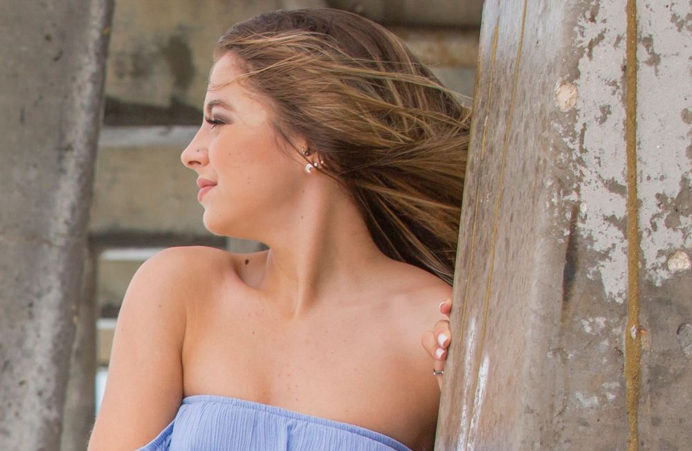 A young woman in a blue off-shoulder top poses thoughtfully against a weathered wooden post.