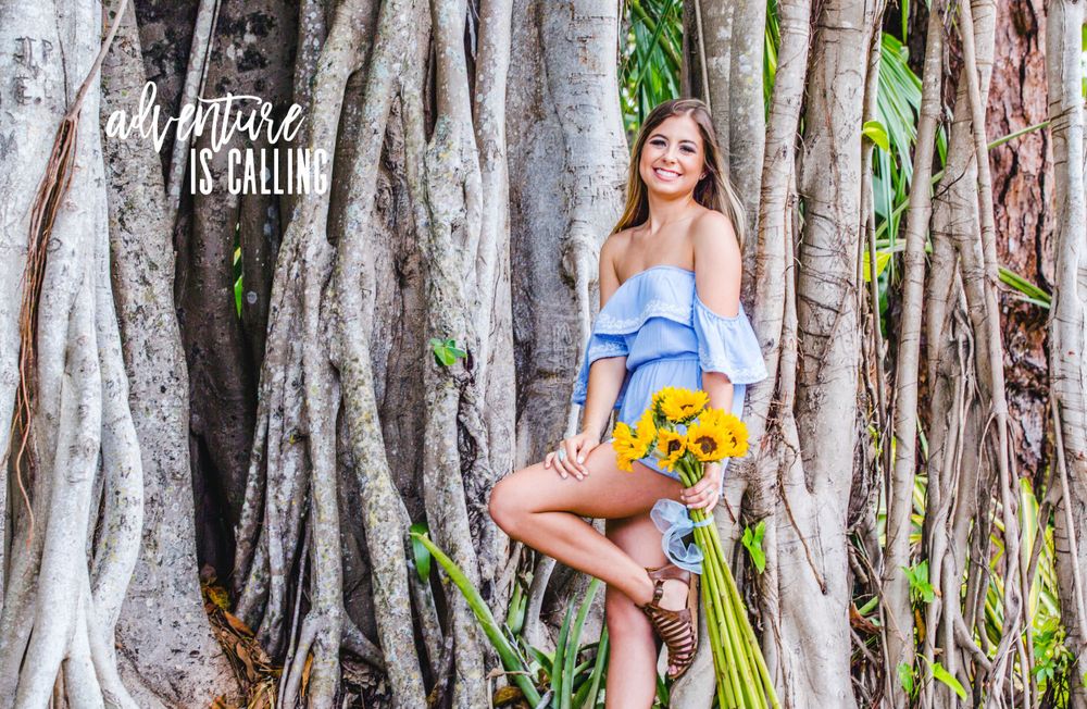 Young woman in blue off-shoulder top holding sunflowers posing among banyan tree roots outdoors.