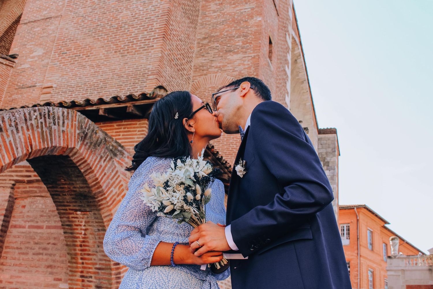 Newlyweds kissing while holding a bouquet against rustic brick architecture.