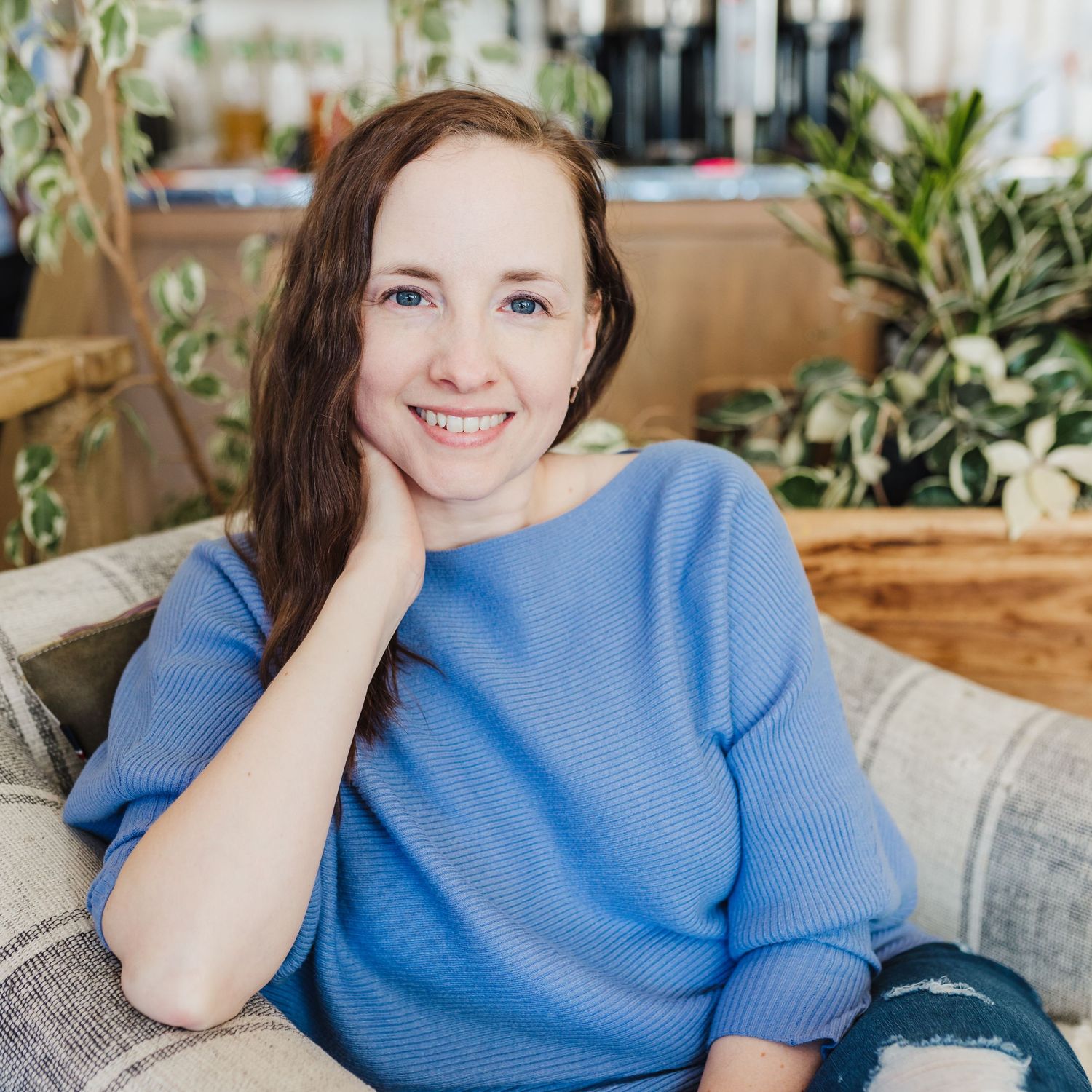 A person in a blue sweater sitting casually on a couch surrounded by houseplants in a bright interior setting.