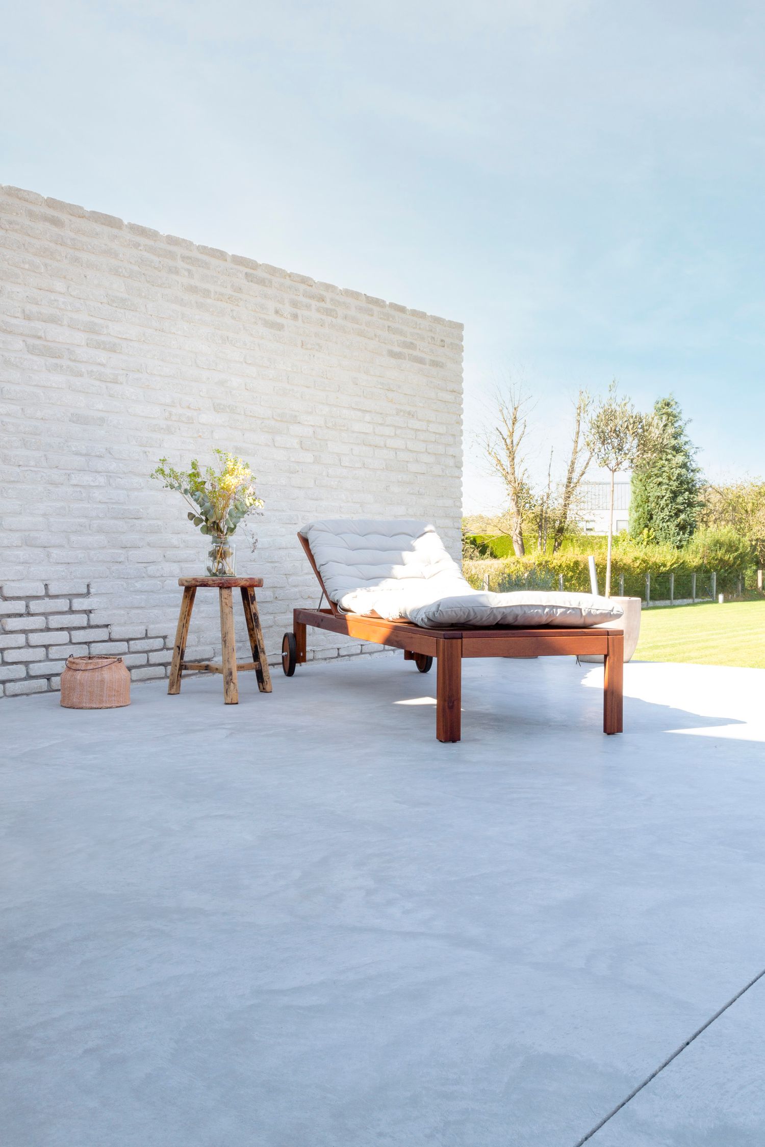 Outdoor patio with wooden lounge chair, white cushions, and floral arrangement against white brick wall.