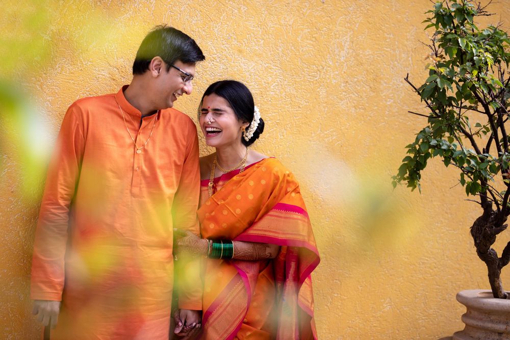 Two people in orange and red traditional Indian attire pose together against a blurred orange backdrop with green plants.