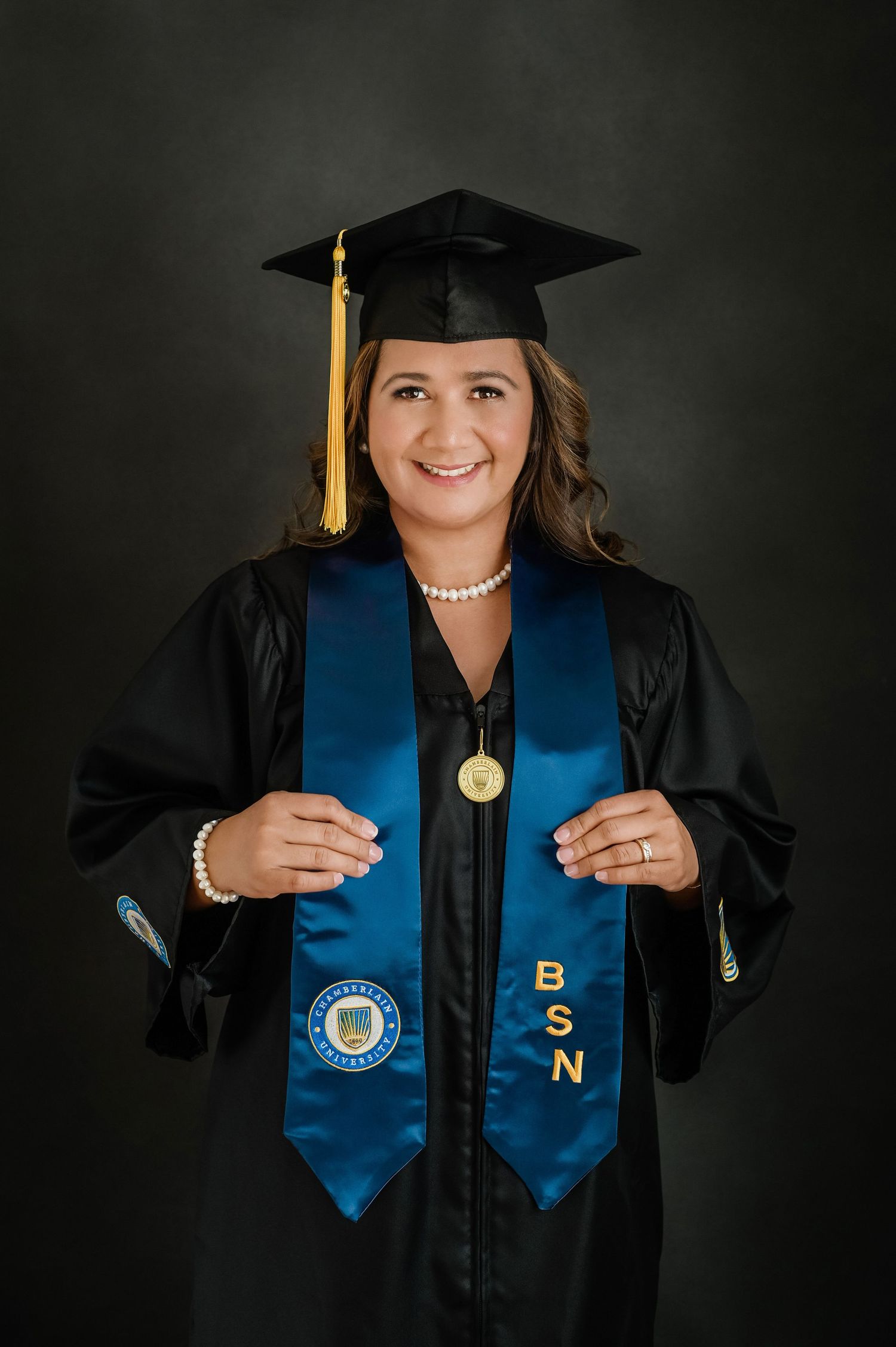 Graduate in cap and gown with blue honors stole and yellow tassel against dark background.