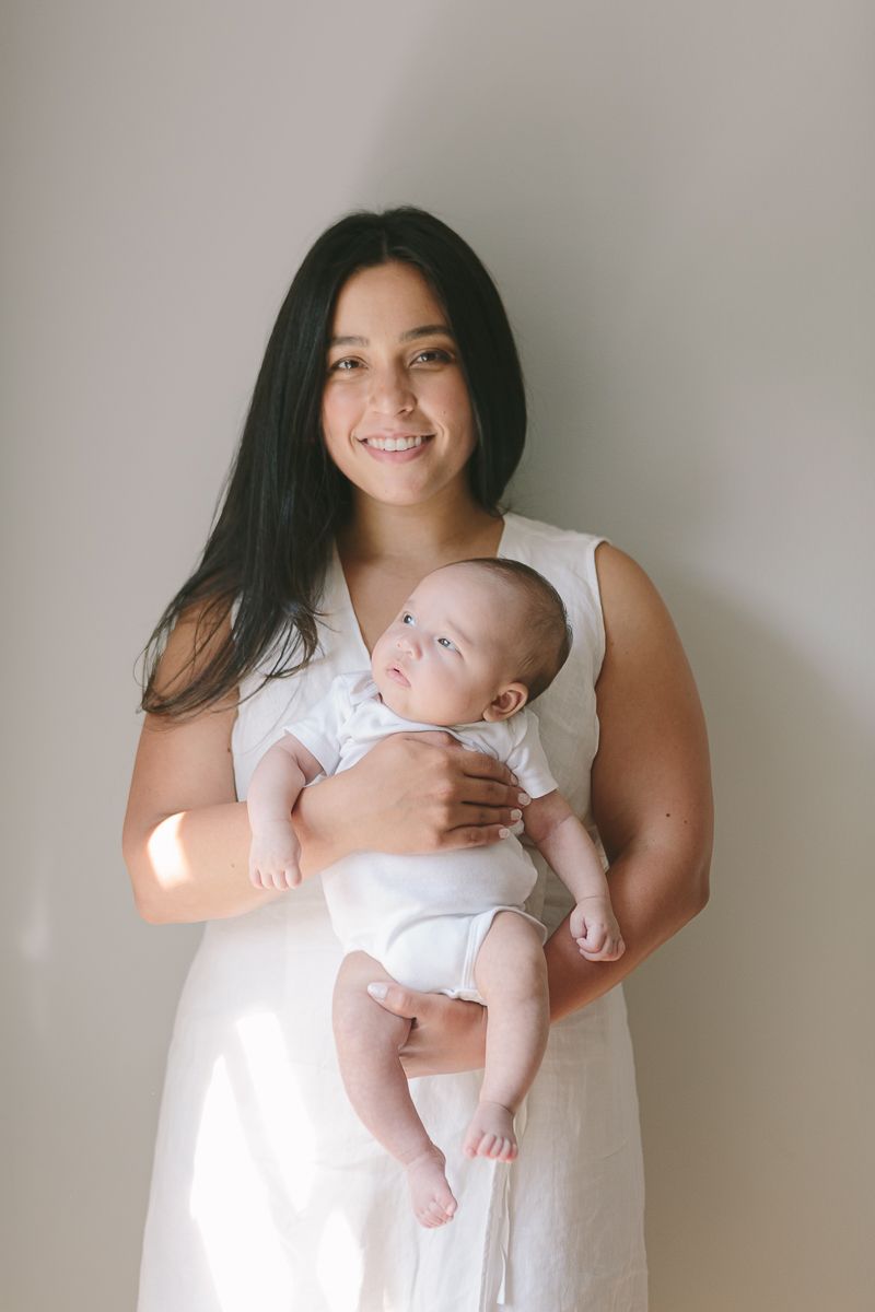 a mother in white dress holding her newborn child on a white background during a newborn session at home in Queens NY