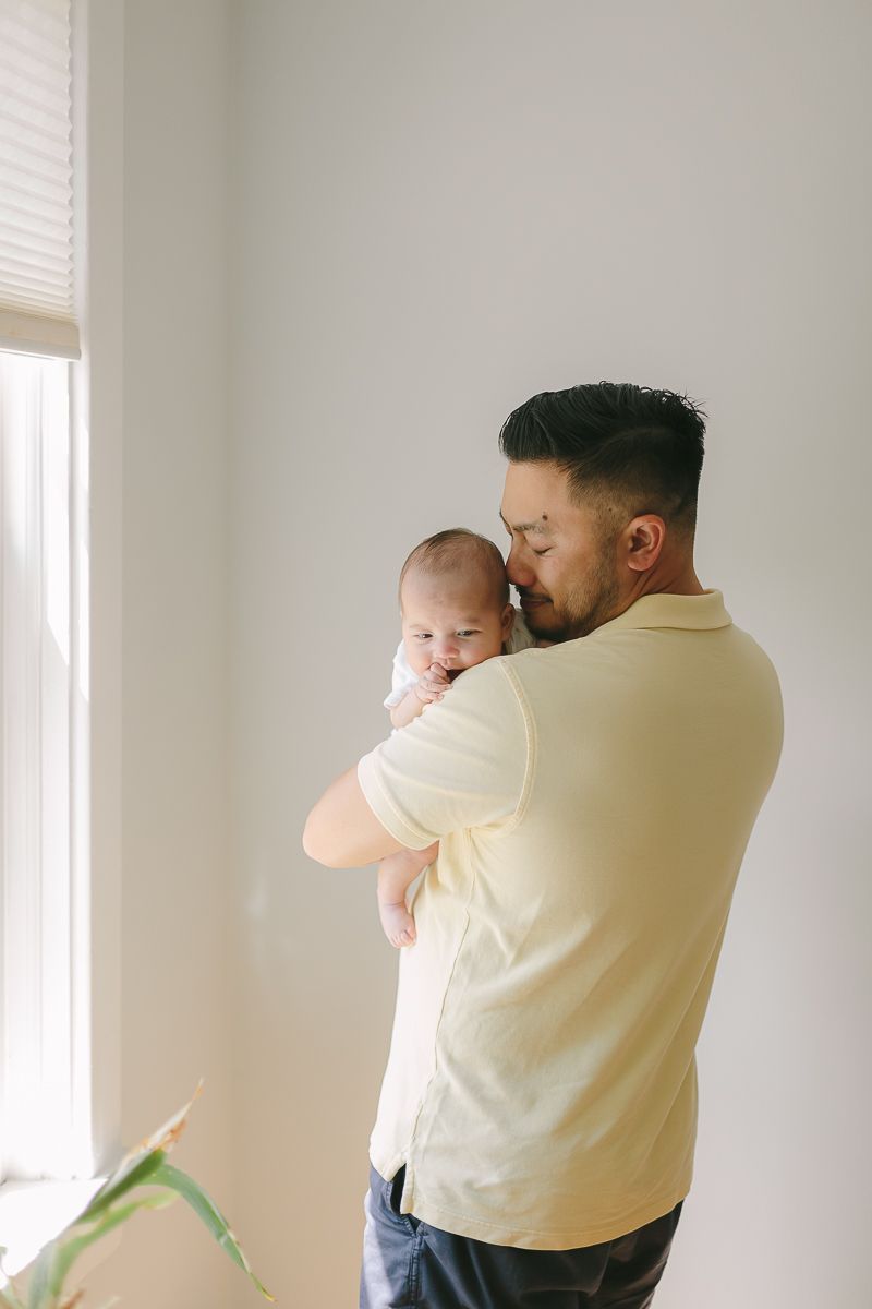 a father wearing a yellow shirt snozzles his newborn baby with beautiful window light during a newborn session in Queens