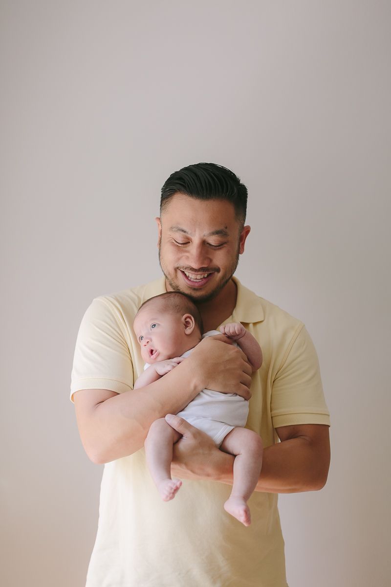 a father wearing a yellow shirt holding his newborn child against hist chest while smiling during a newborn session NYC