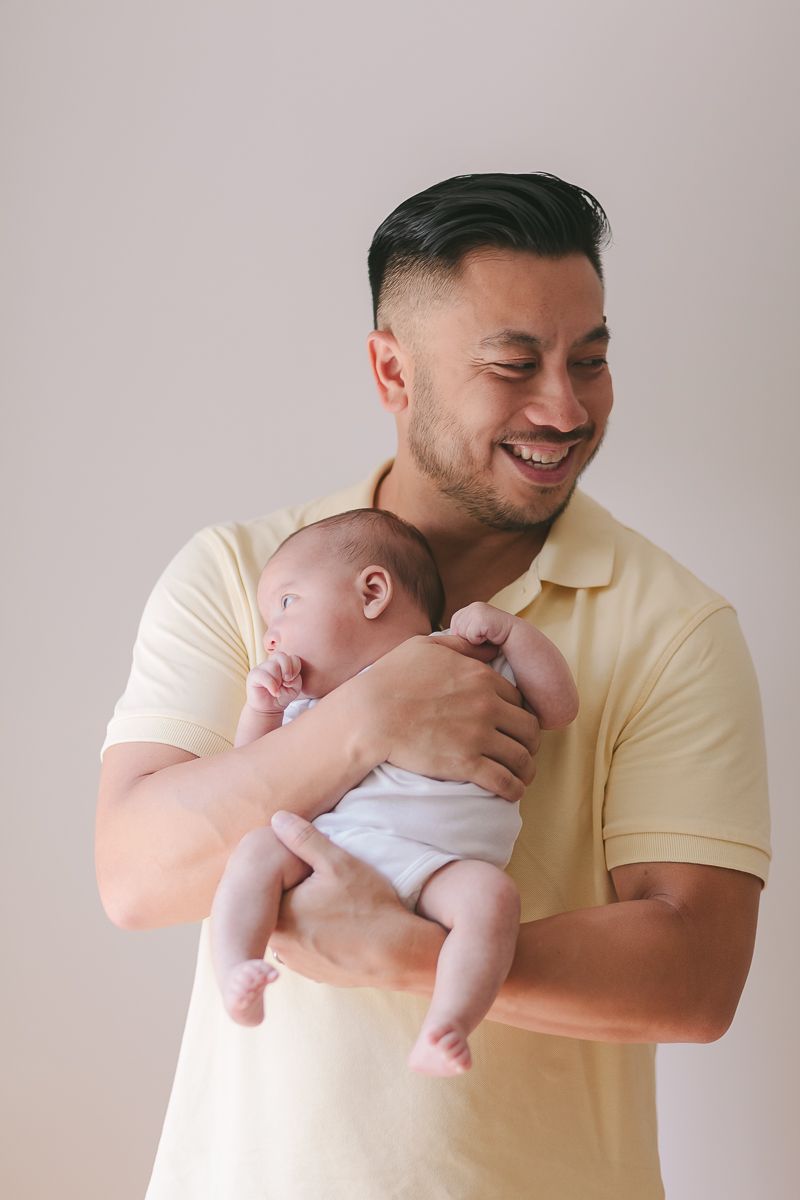 a candid moment of a father smiling while holding his newborn girl on a white background during newborn session at home