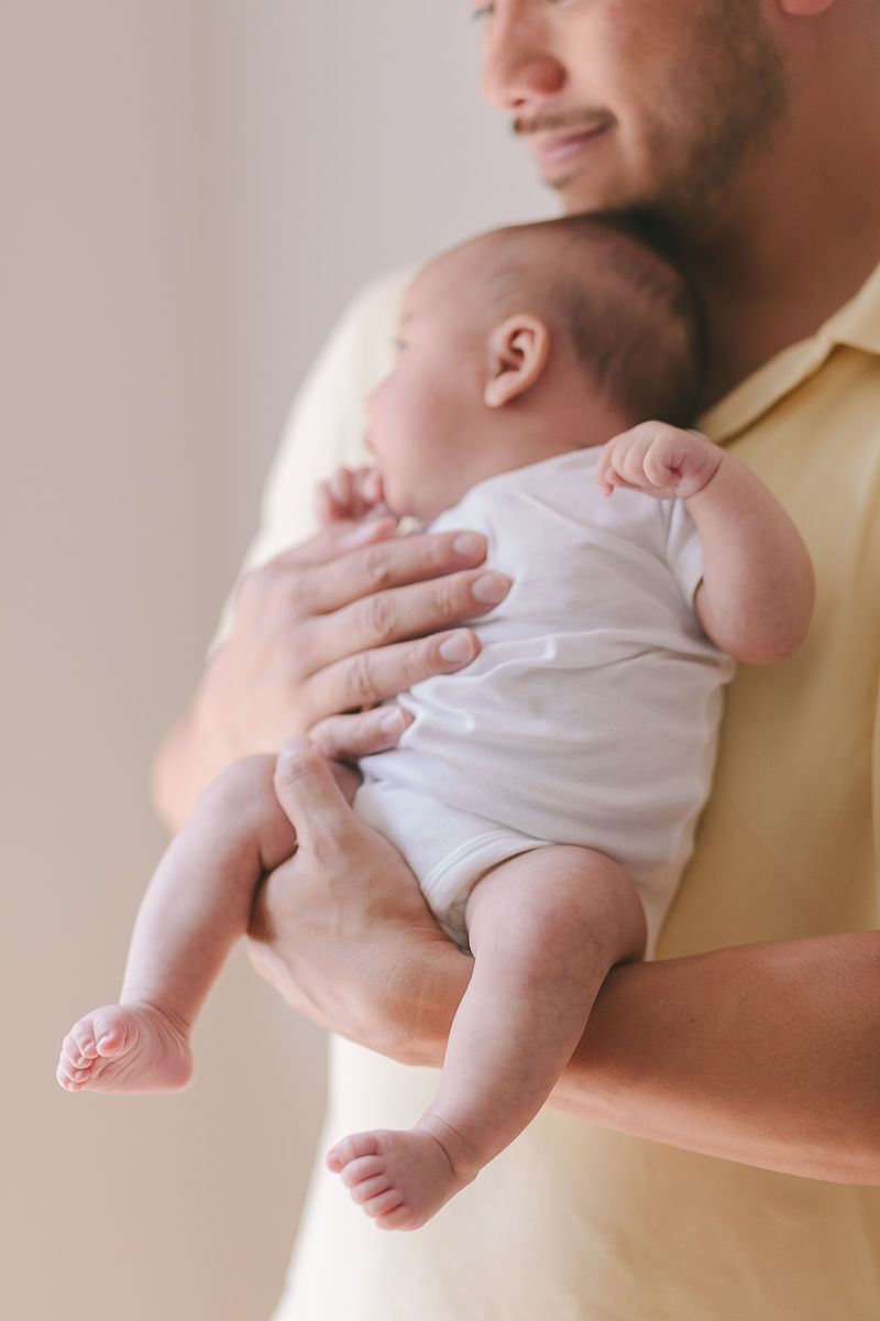 newborn child wearing a white onesie being held by her father photo by Paula Ortiz NYC Family Photographer