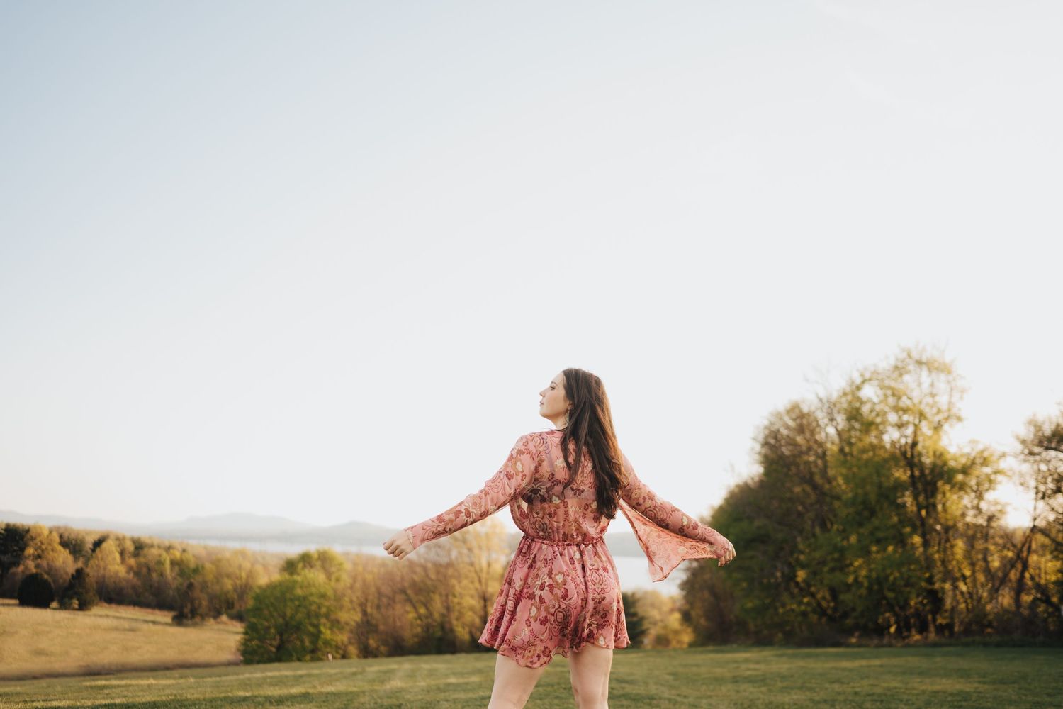 Young woman in a floral pink dress twirls in a golden hour meadow with trees behind her.