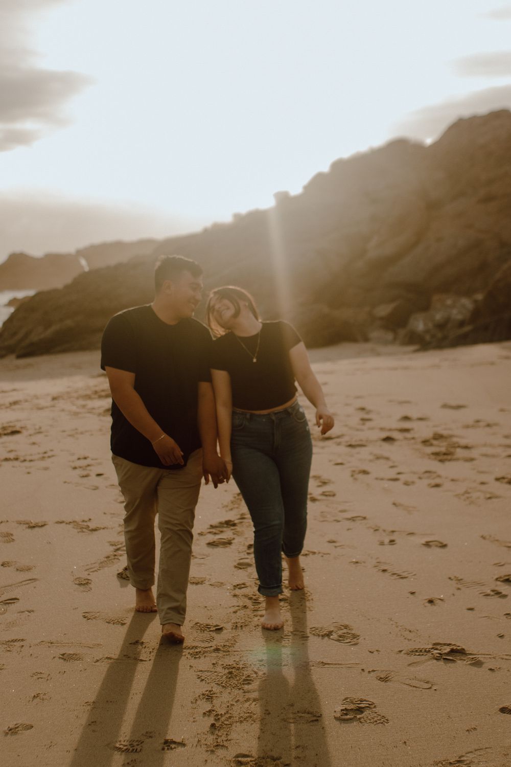 A couple in matching black outfits walking hand in hand on a beach at sunset with cliffs in the background.