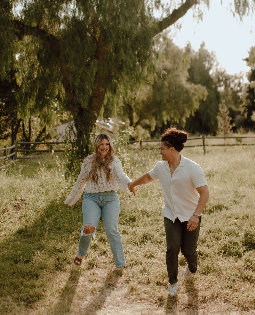 Young couple running and holding hands on a dirt path under willow trees in golden afternoon sunlight.