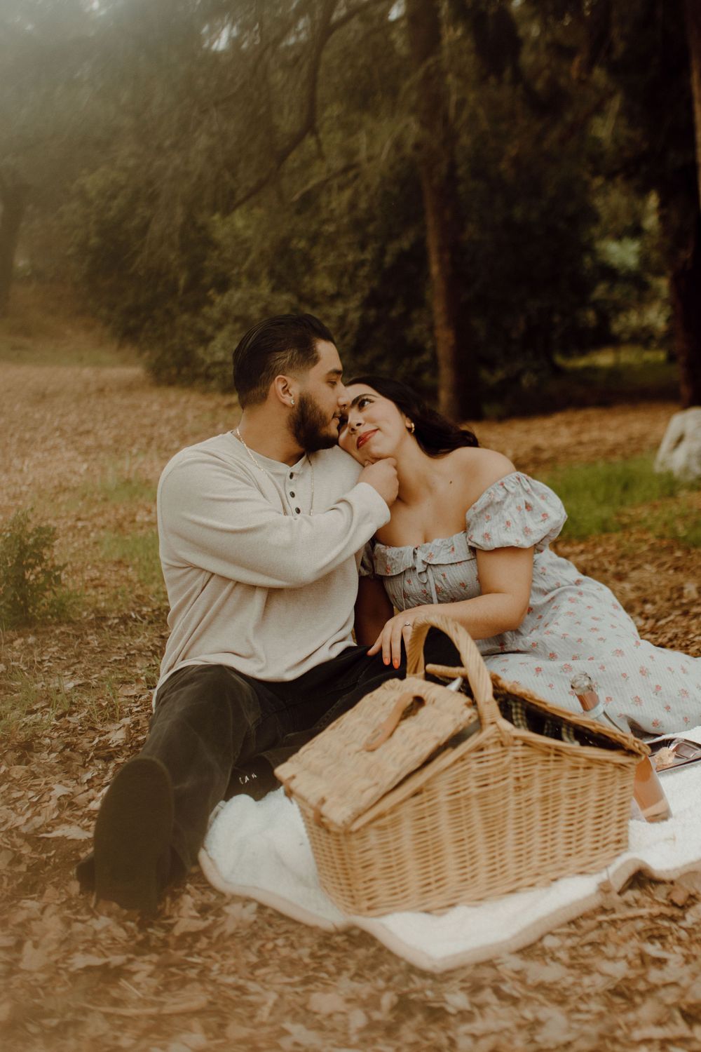 A romantic couple sits on a picnic blanket with a woven basket in a misty wooded setting at sunset.