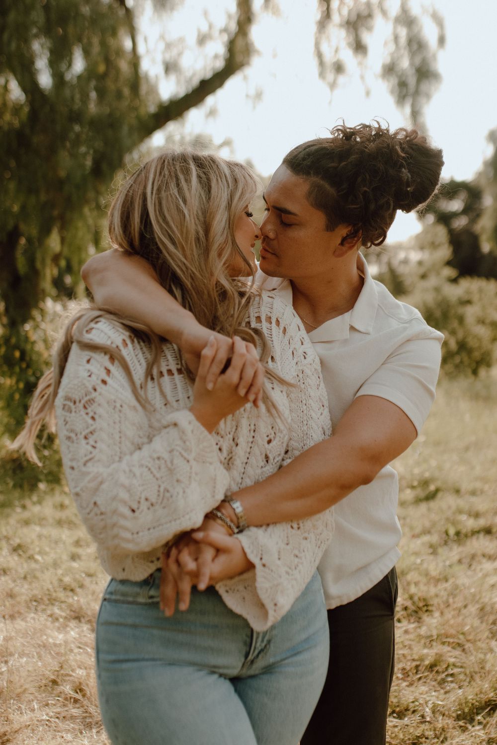 A romantic couple sharing a tender embrace in a sunlit outdoor setting with a willow tree in the background.