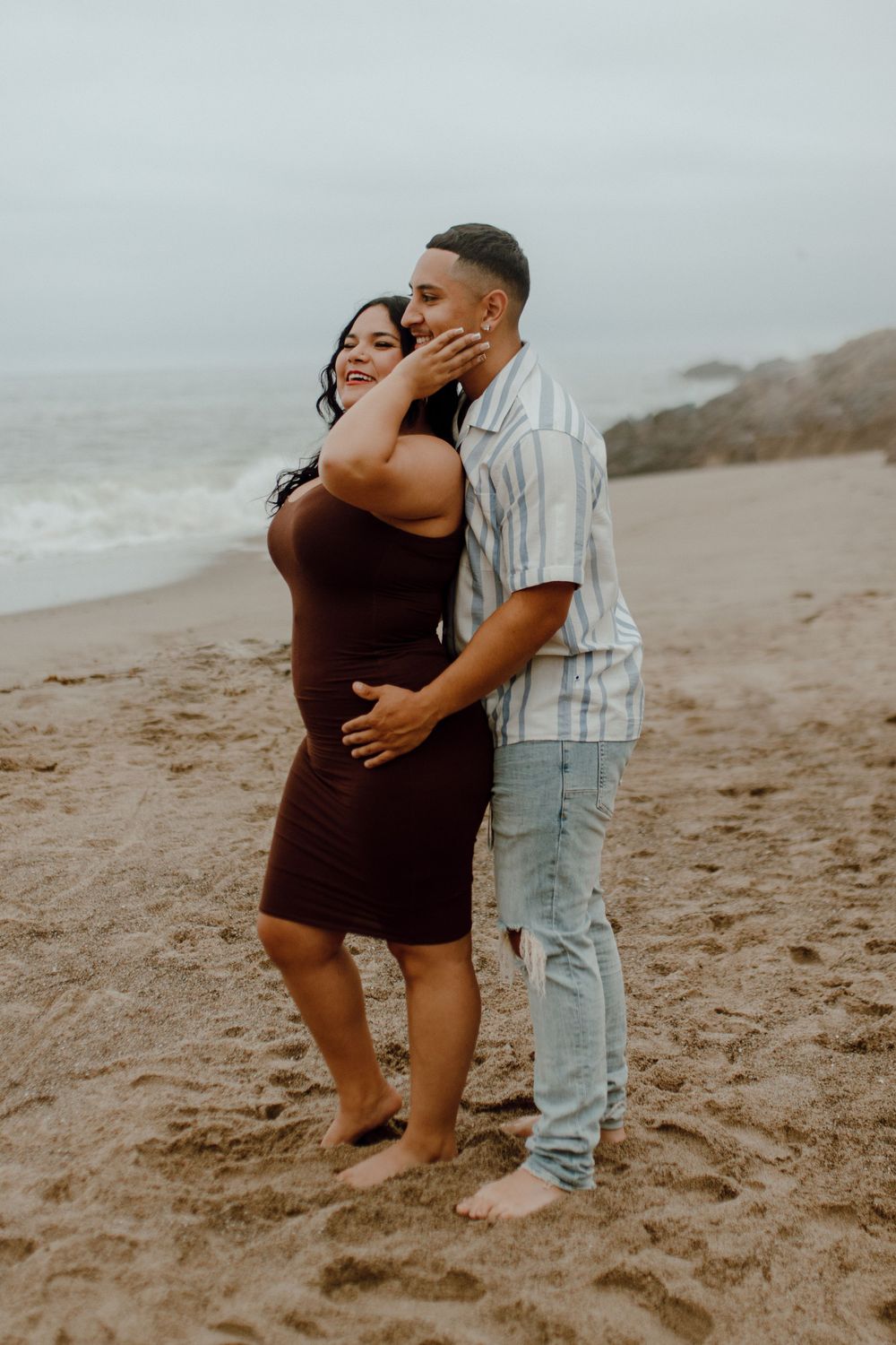 A loving couple embraces on a misty beach shore, sharing a tender moment against the ocean backdrop.