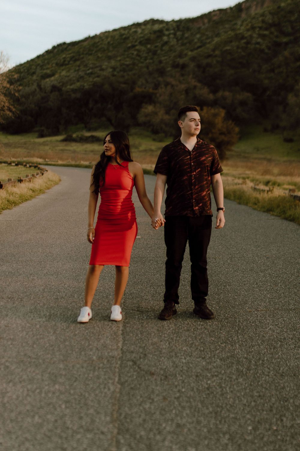 A couple stands on an empty rural road with a green hillside in the background at sunset.