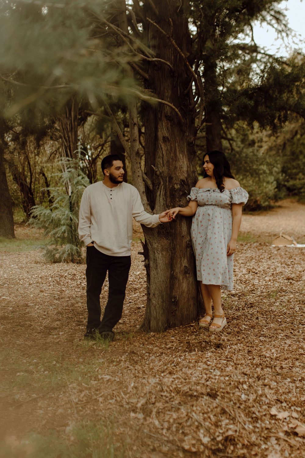 A couple holds hands while standing on opposite sides of a tree trunk in a dreamy outdoor setting at sunset.