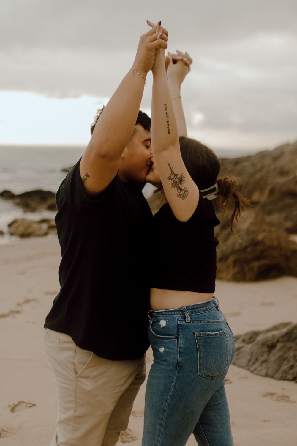 A couple in casual attire dances together on a rocky beach at sunset, with tattoos visible on their raised arms.