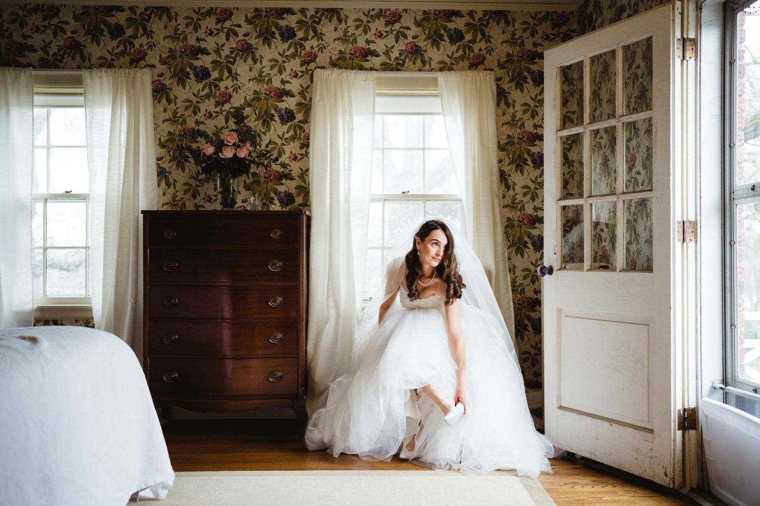 A window-lit room with floral wallpaper features a white wedding dress and wooden dresser.