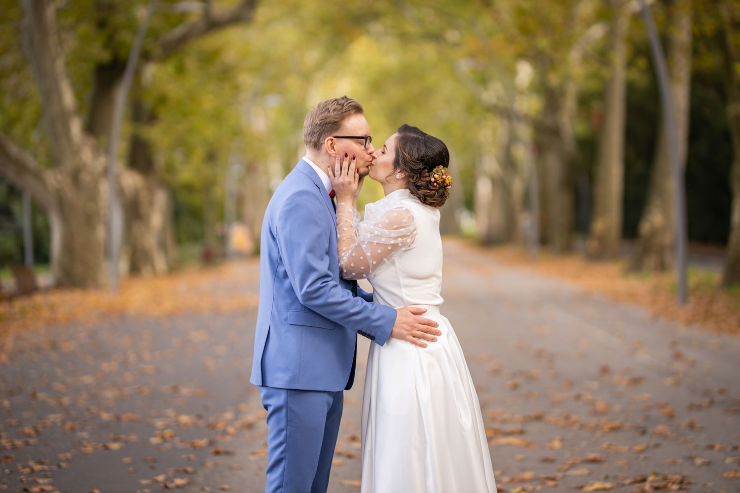 Bride and groom sharing a kiss on a tree-lined autumn path covered in fallen leaves.