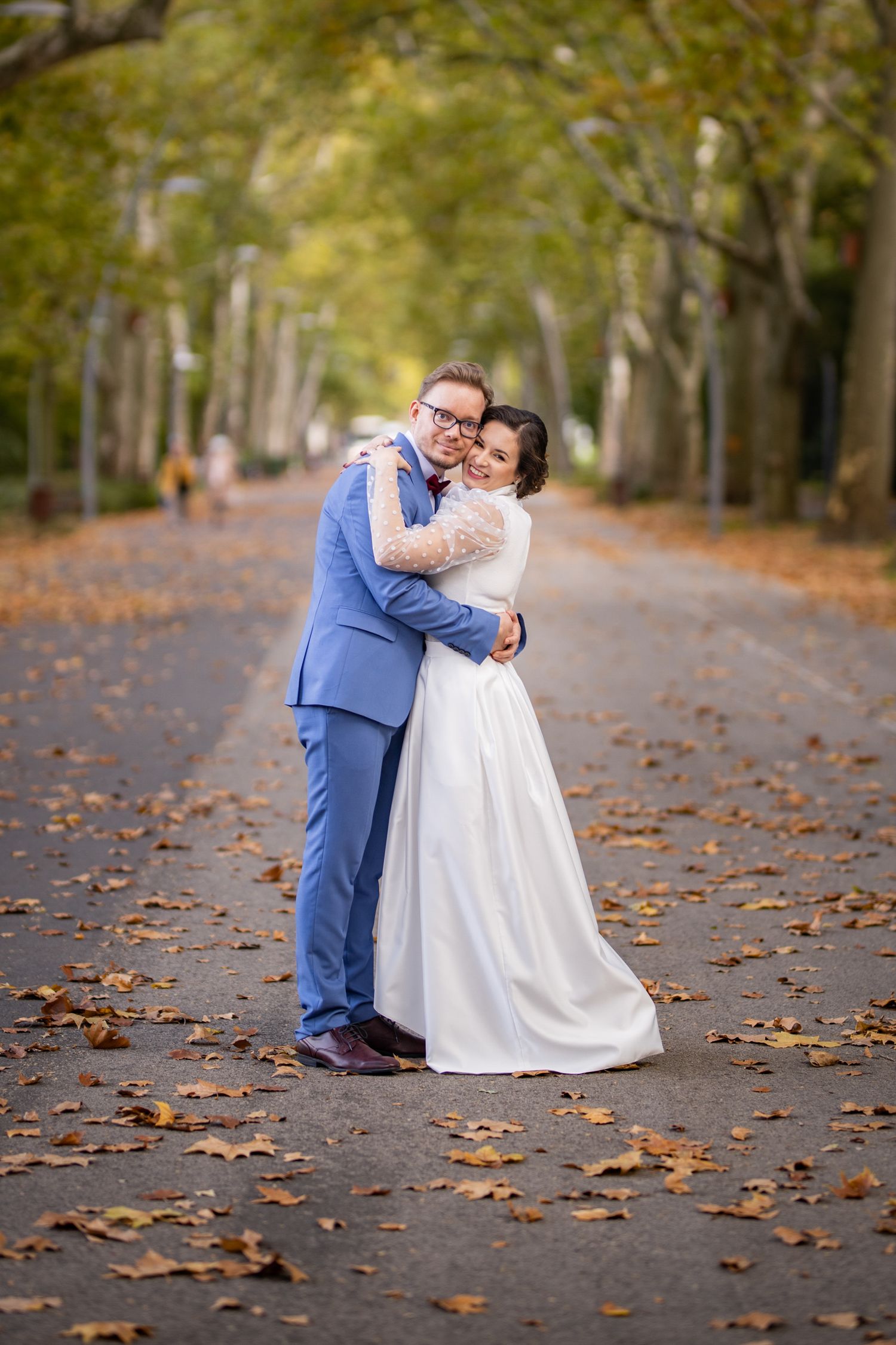 Bride and groom embracing on a scenic autumn avenue with golden fallen leaves.