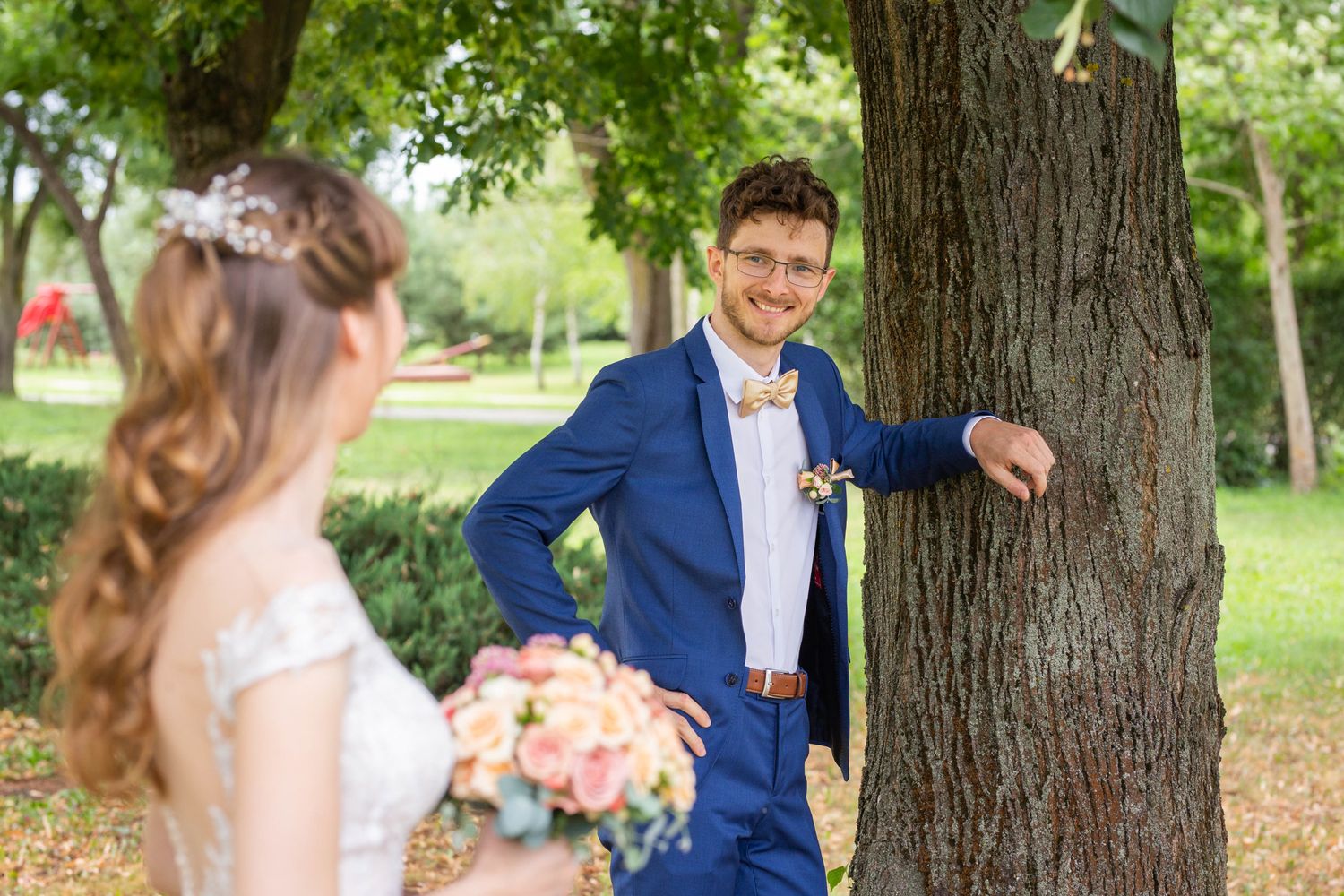 Groom in blue suit smiling at bride from behind a tree in a sunny park setting.