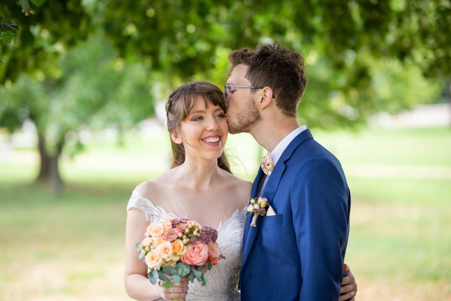 Groom in blue suit kissing bride on cheek as she smiles holding a peach bouquet.
