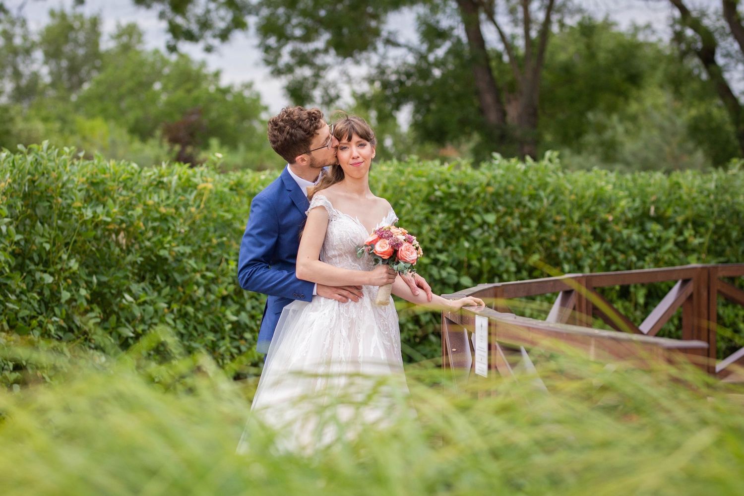 Groom in blue suit kissing bride on cheek as she holds a pink bouquet on a lush green bridge.
