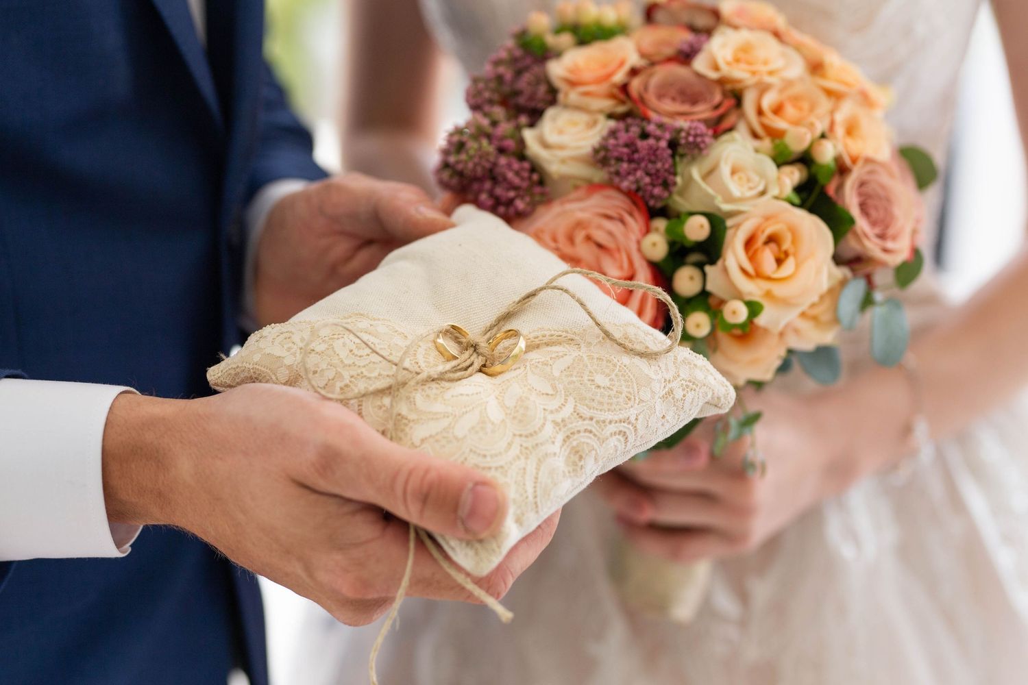 Close-up of wedding rings on an ornate pillow held by groom beside bride holding a peach floral bouquet.