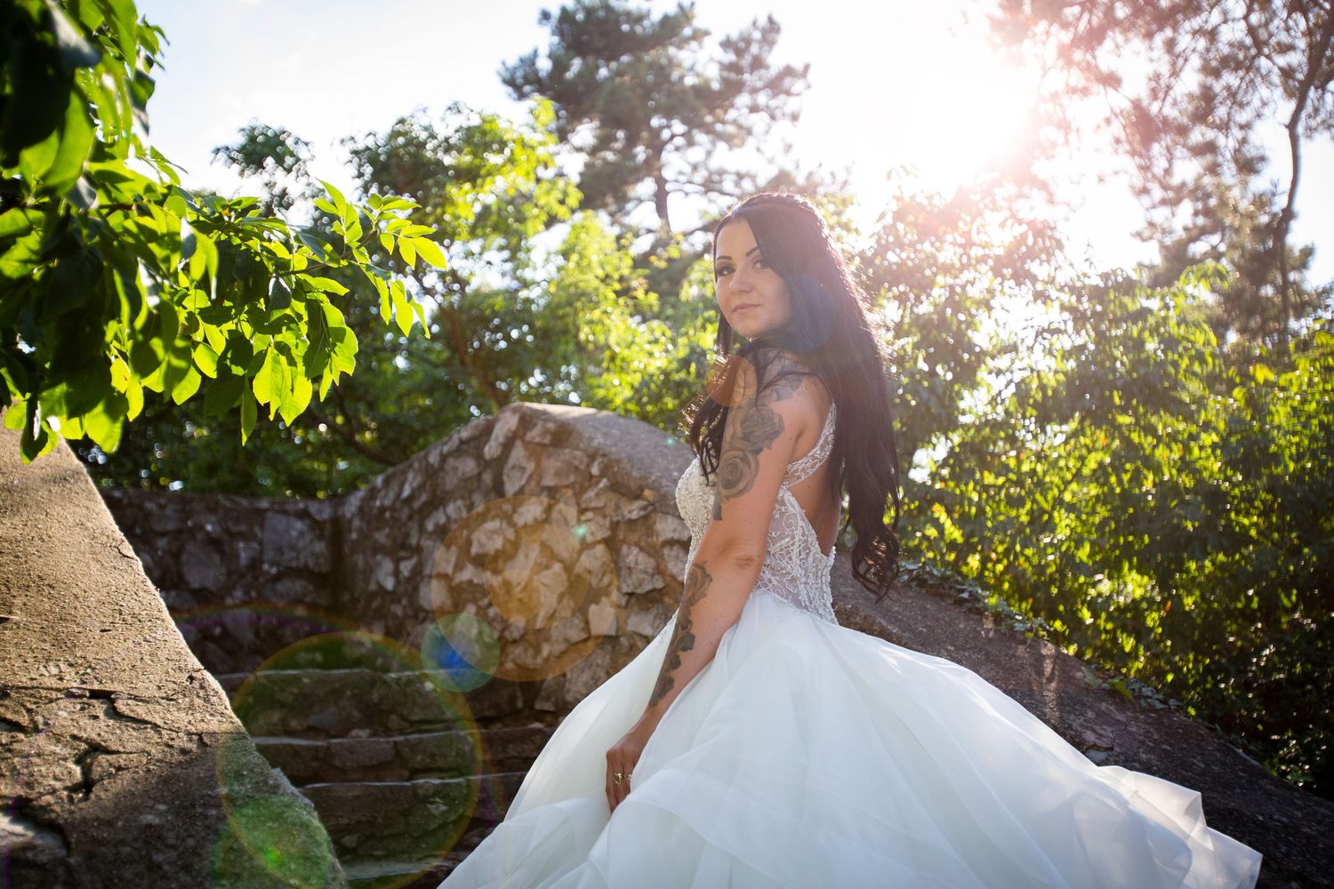 Bride in a voluminous white ball gown posing in a sunlit garden with stone walls.
