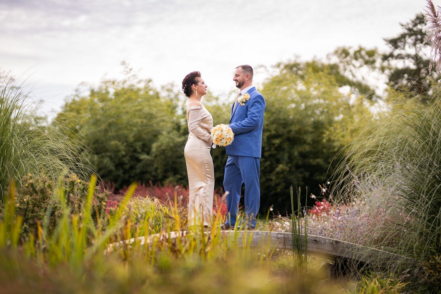 Bride and groom in a colorful garden, facing each other lovingly, groom in blue suit holding bouquet.
