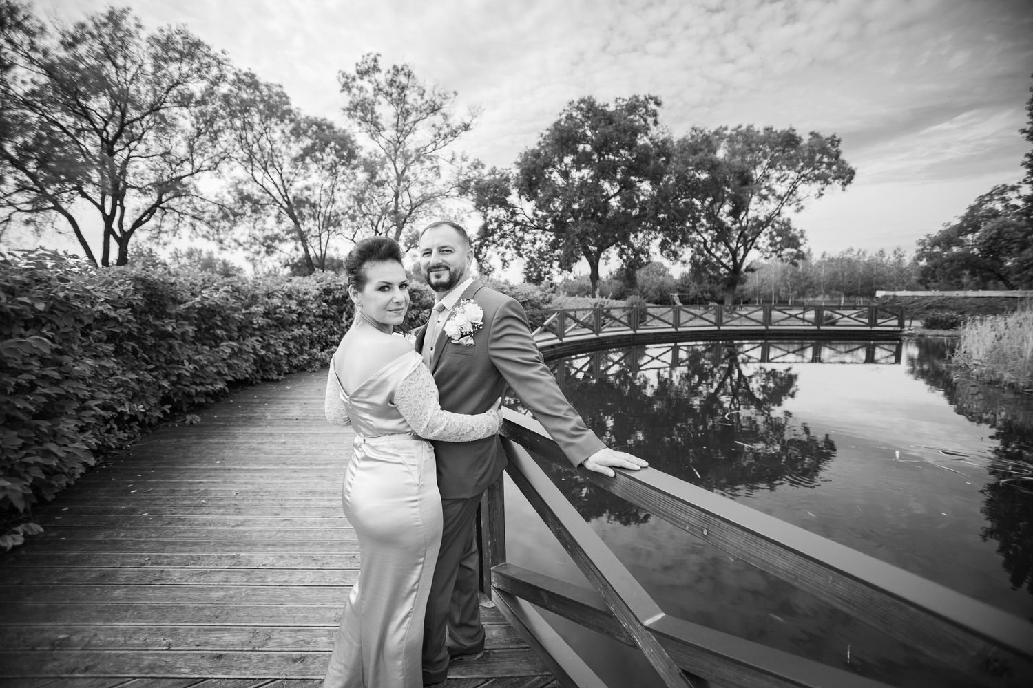 Black and white photo of bride and groom embracing on a wooden bridge over a calm pond.