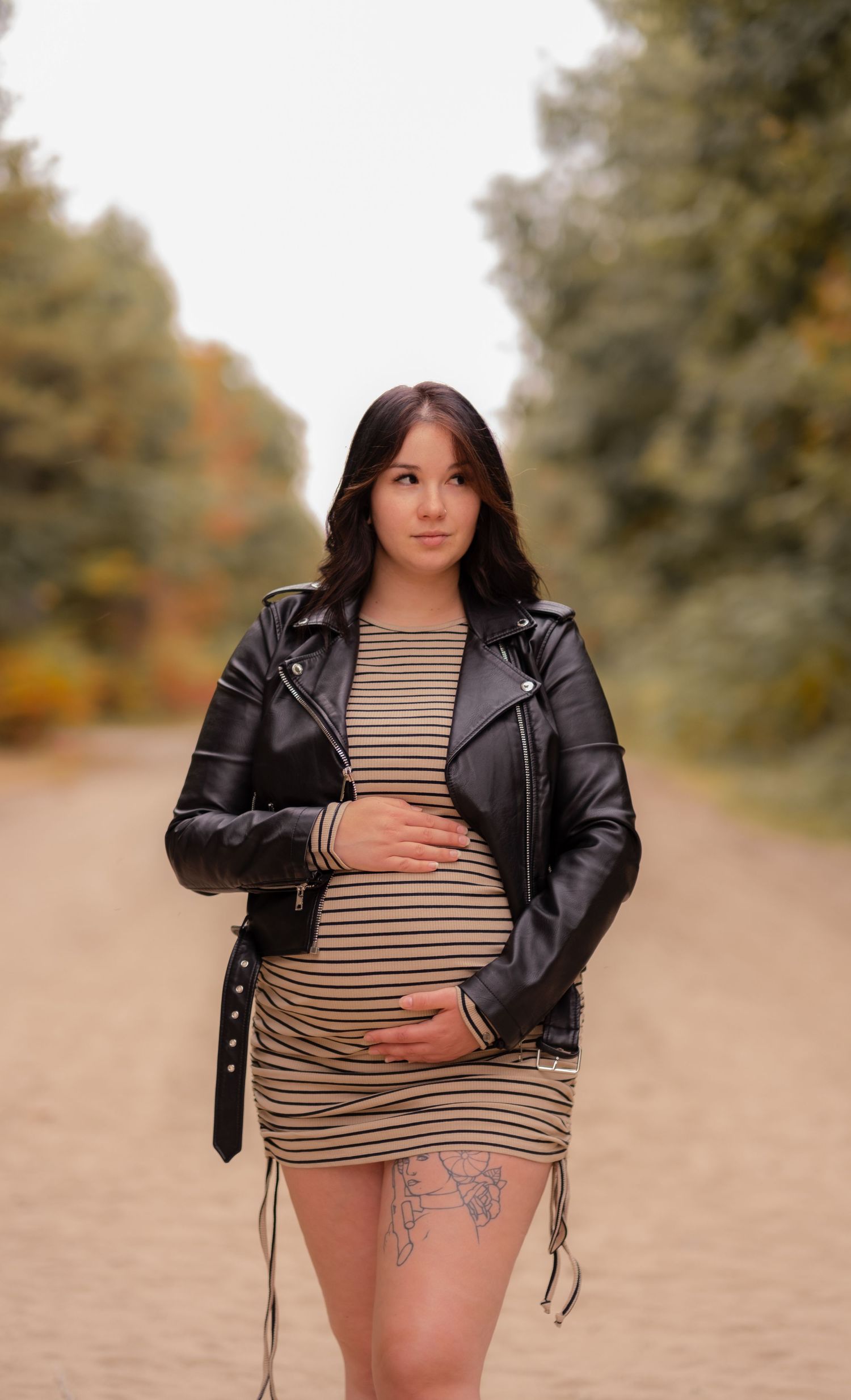 Expecting person in striped dress and leather jacket poses on autumn path.