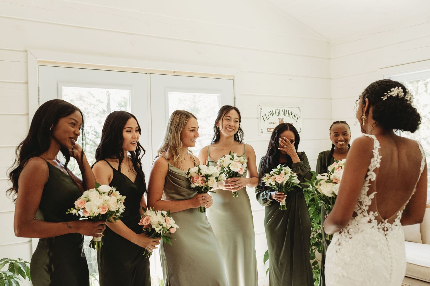 Bridesmaids in black dresses hold white bouquets while getting ready in a bright room.
