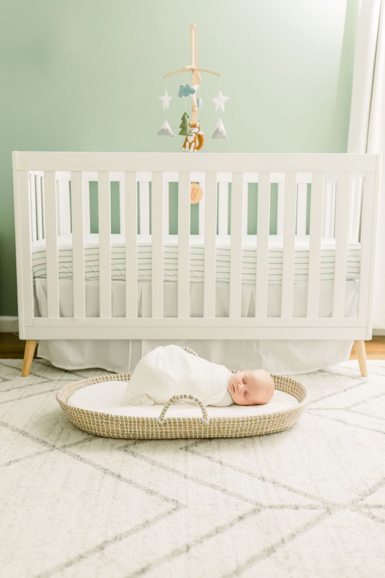 White modern crib with mobile and sleeping infant in basket on geometric rug below.