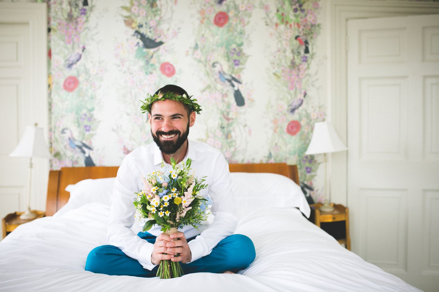 A person in white sits cross-legged on a bed holding flowers with floral wallpaper behind.