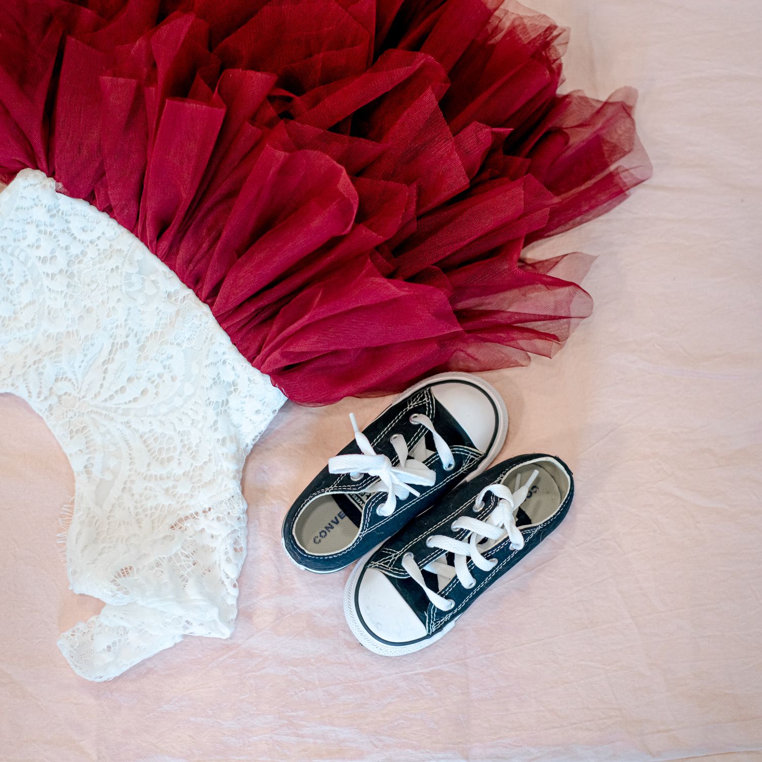 Black Converse sneakers next to red tulle and white lace dress on wooden floor.