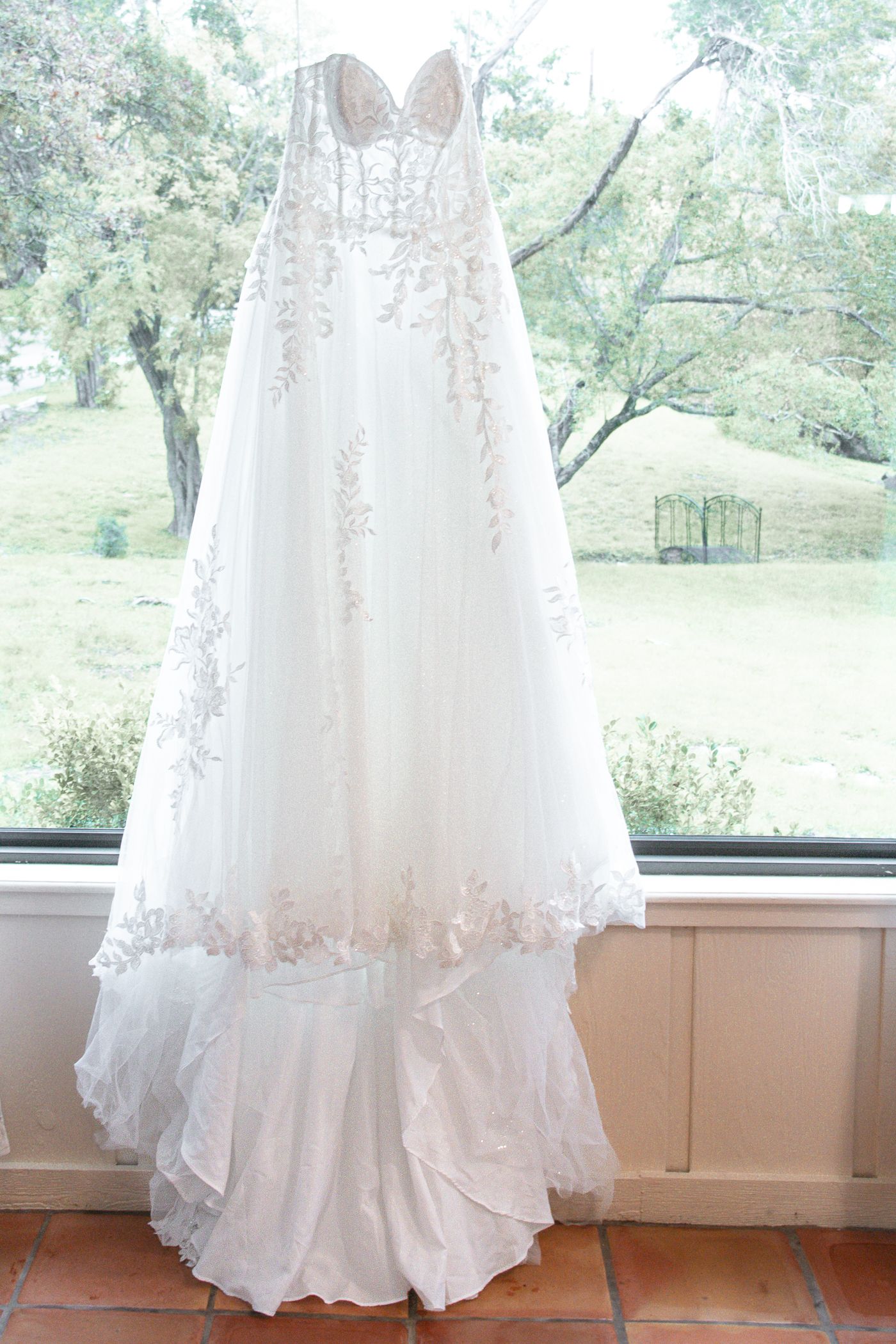 An elegant white lace wedding dress hangs by a window overlooking a scenic green garden.