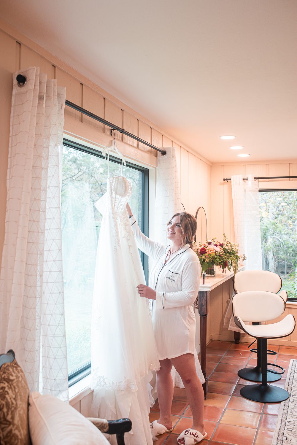 A wedding dress hangs by a window in a light-filled room while someone in white attends to it.