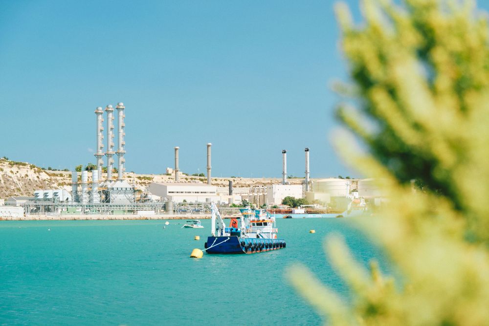 Industrial port with smokestacks and blue cargo ship in crystal clear turquoise waters.