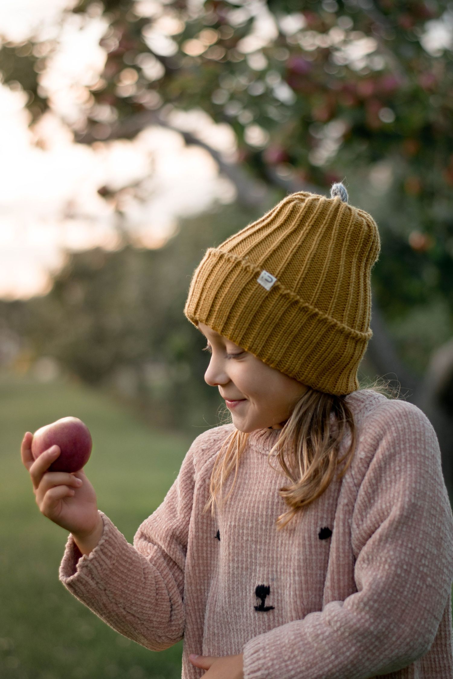 A person in a pink sweater examines a fresh apple while wearing a mustard knit beanie.
