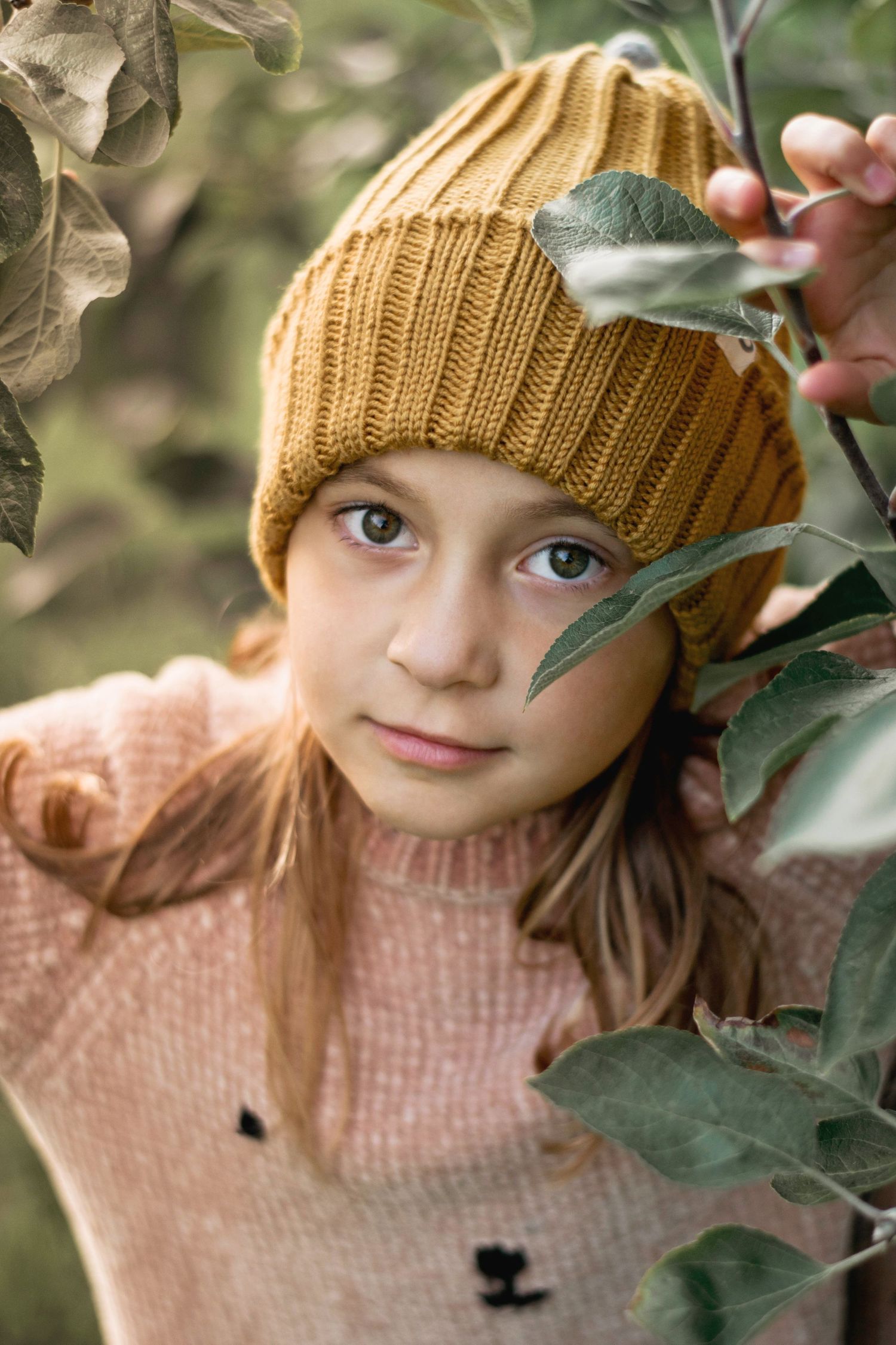 A close-up nature portrait featuring mustard beanie and pink sweater among apple tree leaves.