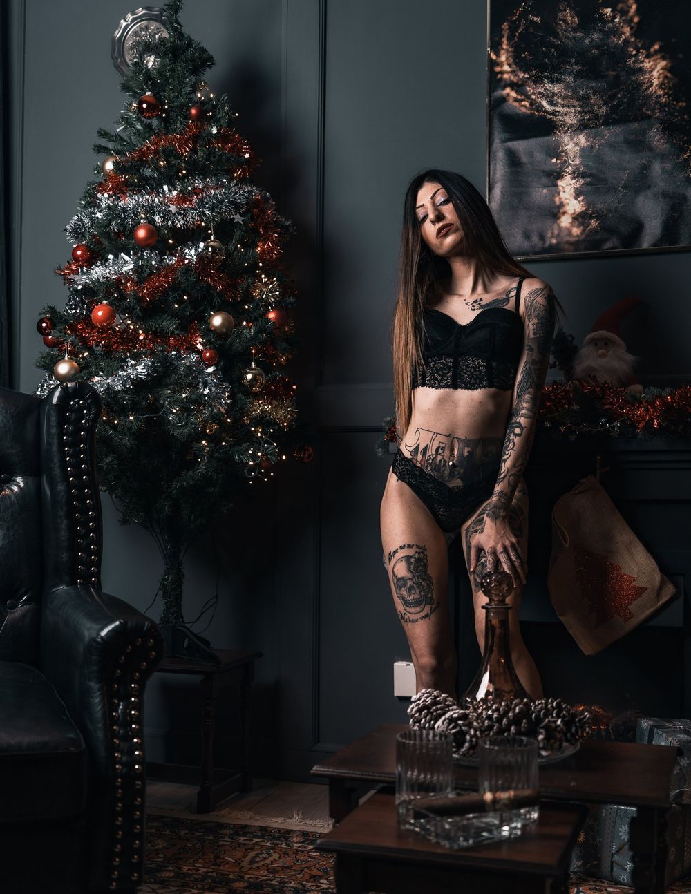Dark moody Christmas scene with decorated tree and tattooed model in black lingerie.
