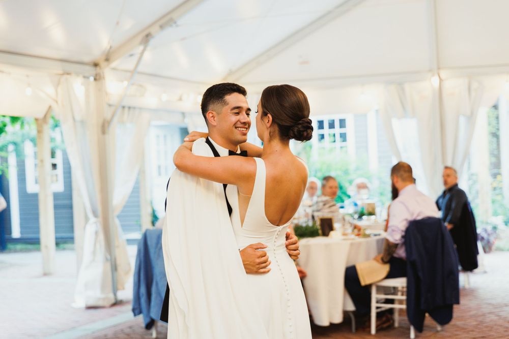 Newlyweds share their first dance in an elegant white tent reception venue with guests looking on.