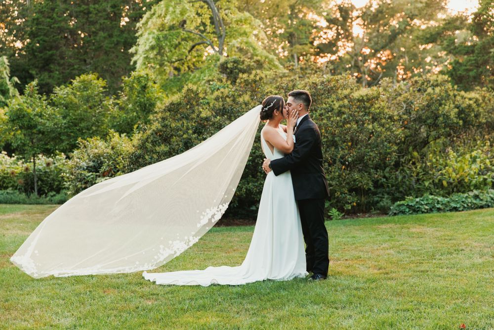 Wedding couple embracing on green lawn with dramatic flowing veil in summer garden.